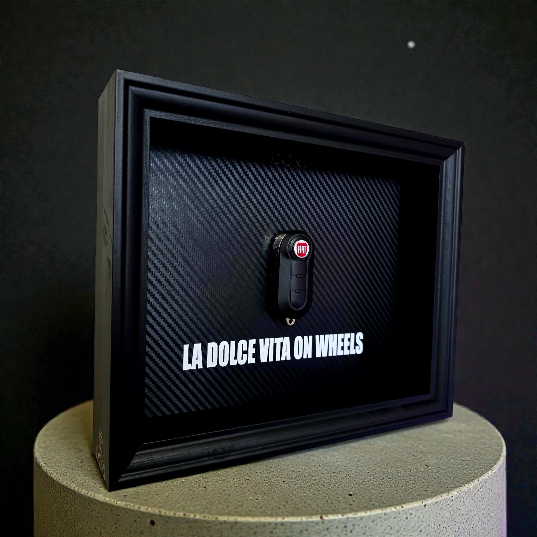 Automobilio raktelio dekoratyvinis paveikslas su anglies pluošto fonu ir užrašu „La Dolce Vita on Wheels“