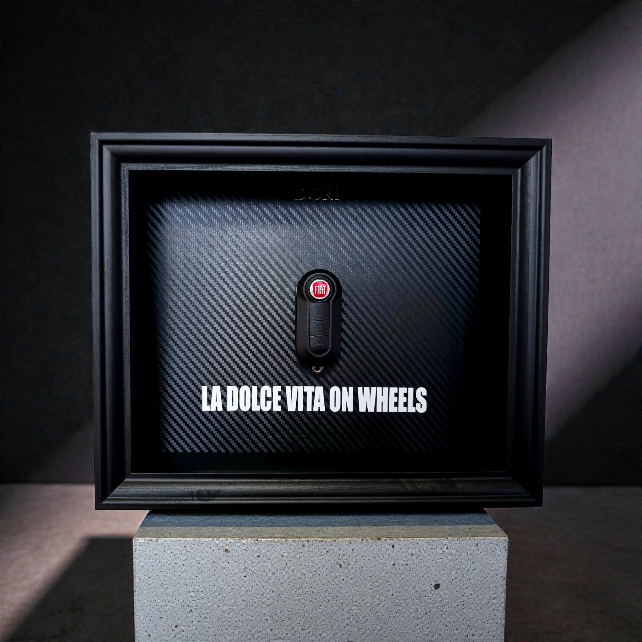 Automobilio raktelio dekoratyvinis paveikslas su anglies pluošto fonu ir užrašu „La Dolce Vita on Wheels“