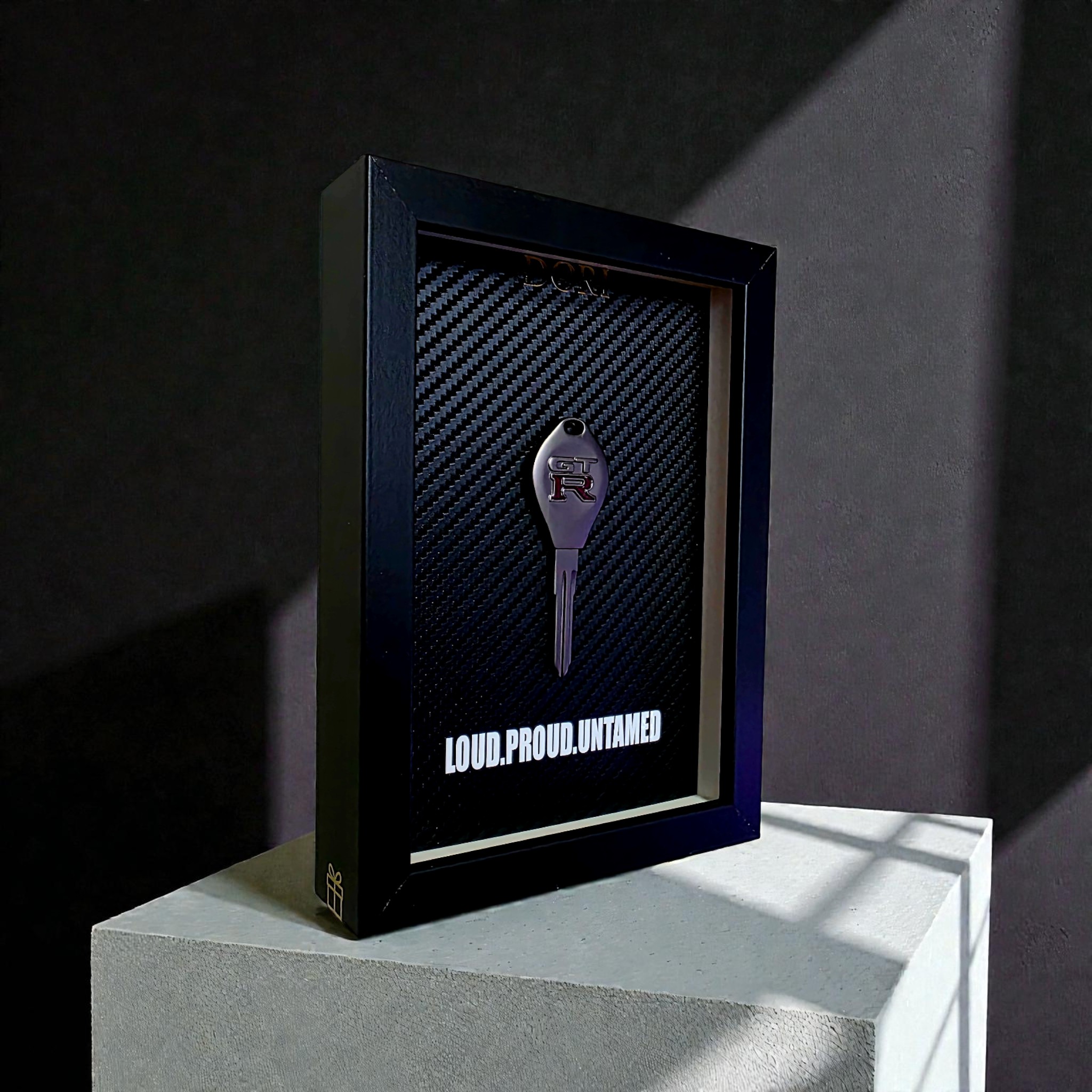 Automobilio raktelio dekoratyvinis paveikslas su anglies pluošto fonu ir užrašu „Loud. Proud. Untamed.“