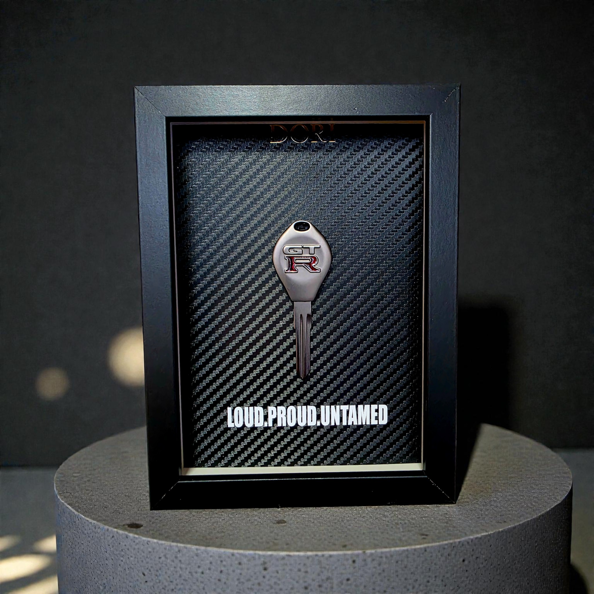 Automobilio raktelio dekoratyvinis paveikslas su anglies pluošto fonu ir užrašu „Loud. Proud. Untamed.“