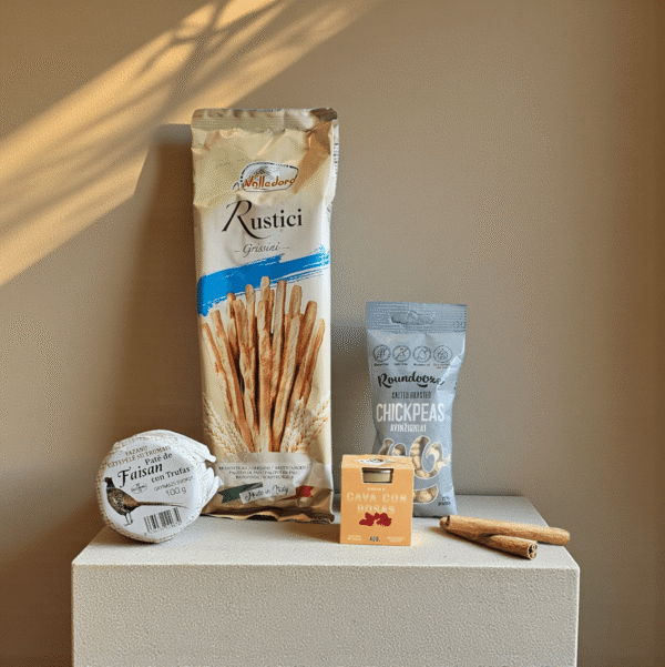 DORI gurmaniškas rinkinys su grissini lazdelėmis, skrudintais avinžirniais, paštetu ir sausainiais, fotografuotas ant šilto smėlio fono.