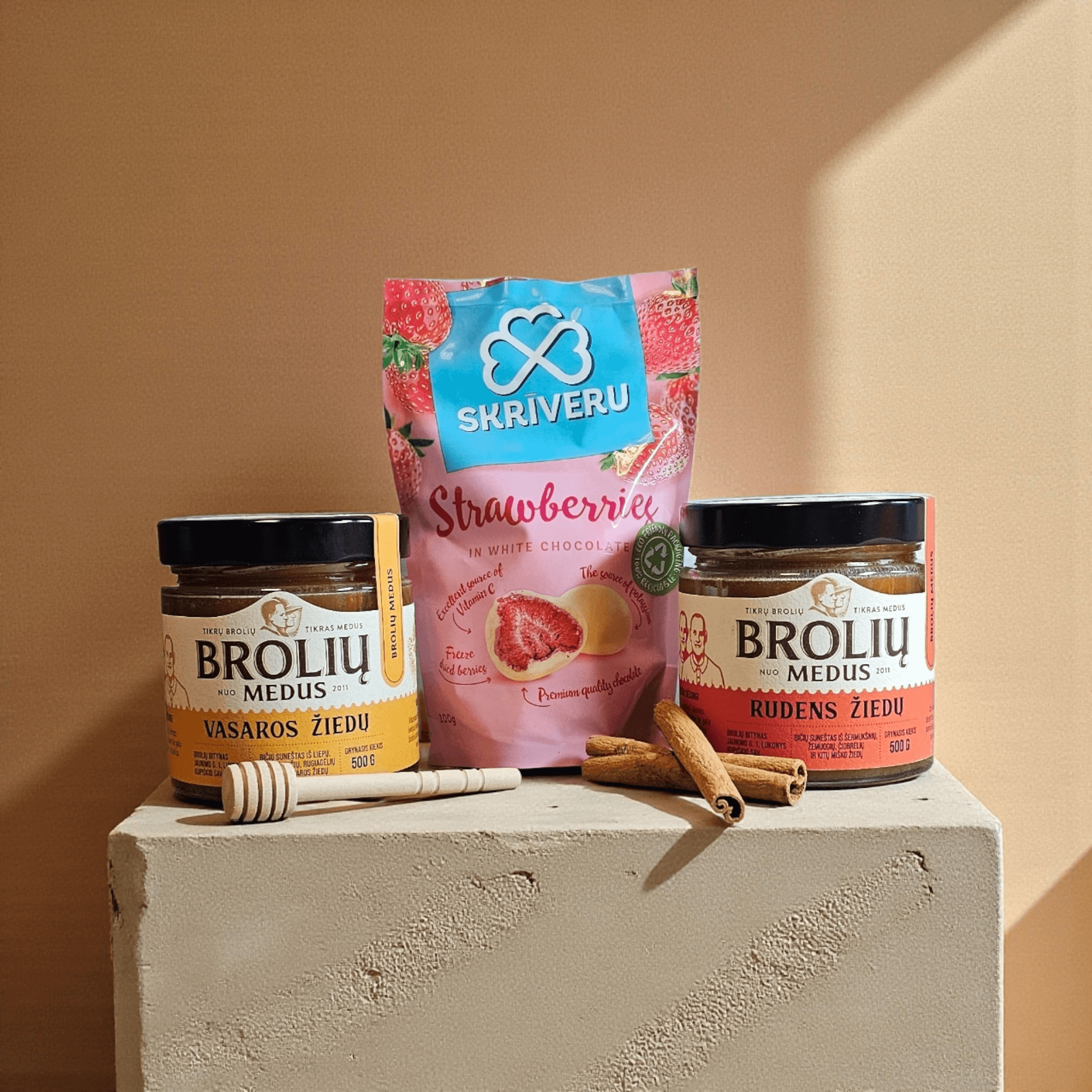 DORI saldus rinkinys su vasaros ir rudens žiedų medumi, braškėmis baltame šokolade ir cinamono lazdelėmis, fotografuotas šiltame minimalistiniame fone.