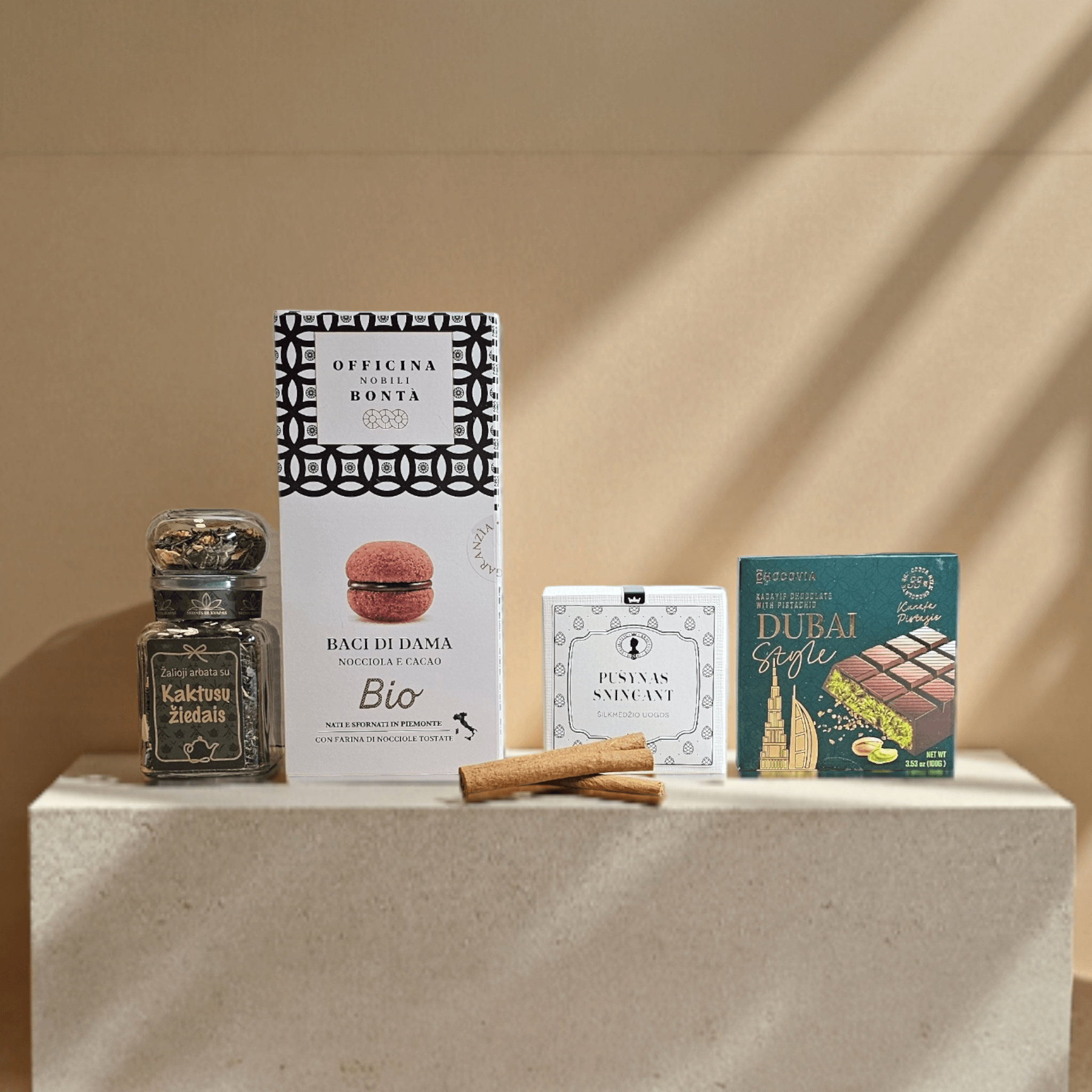 DORI gurmaniškas rinkinys su kakavos arbata, itališkais Baci di Dama sausainiais, premium plytelių šokoladu „Dubai“ ir elegantišku sausainiu, fotografuotas šiltame minimalistiniame fone.
