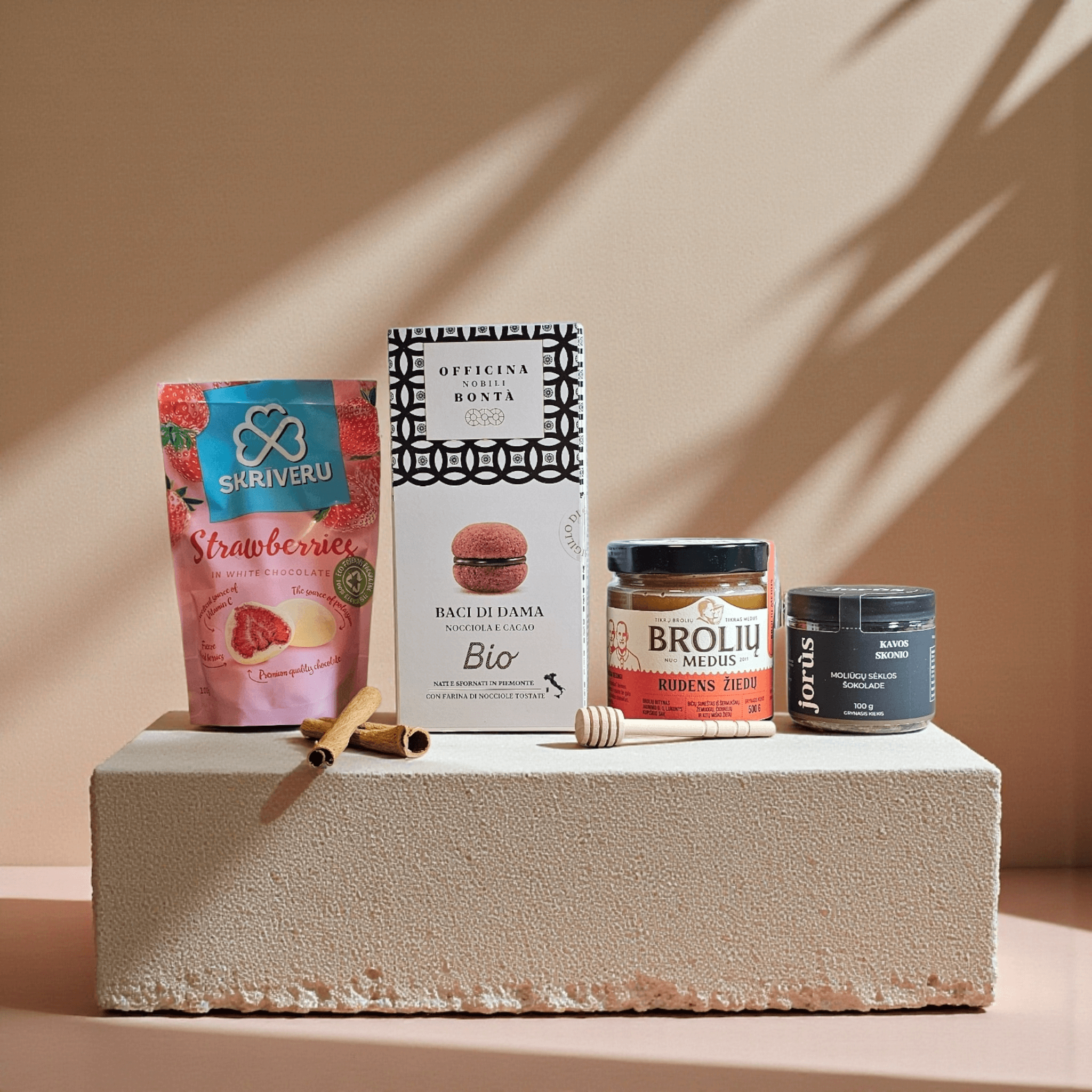 DORI gurmaniškas rinkinys su braškėmis baltame šokolade, itališkais Baci di Dama sausainiais, Brolių rudens žiedų medumi ir kivio skonio arbata, fotografuotas šiltame minimalistiniame fone su cinamono lazdelėmis.