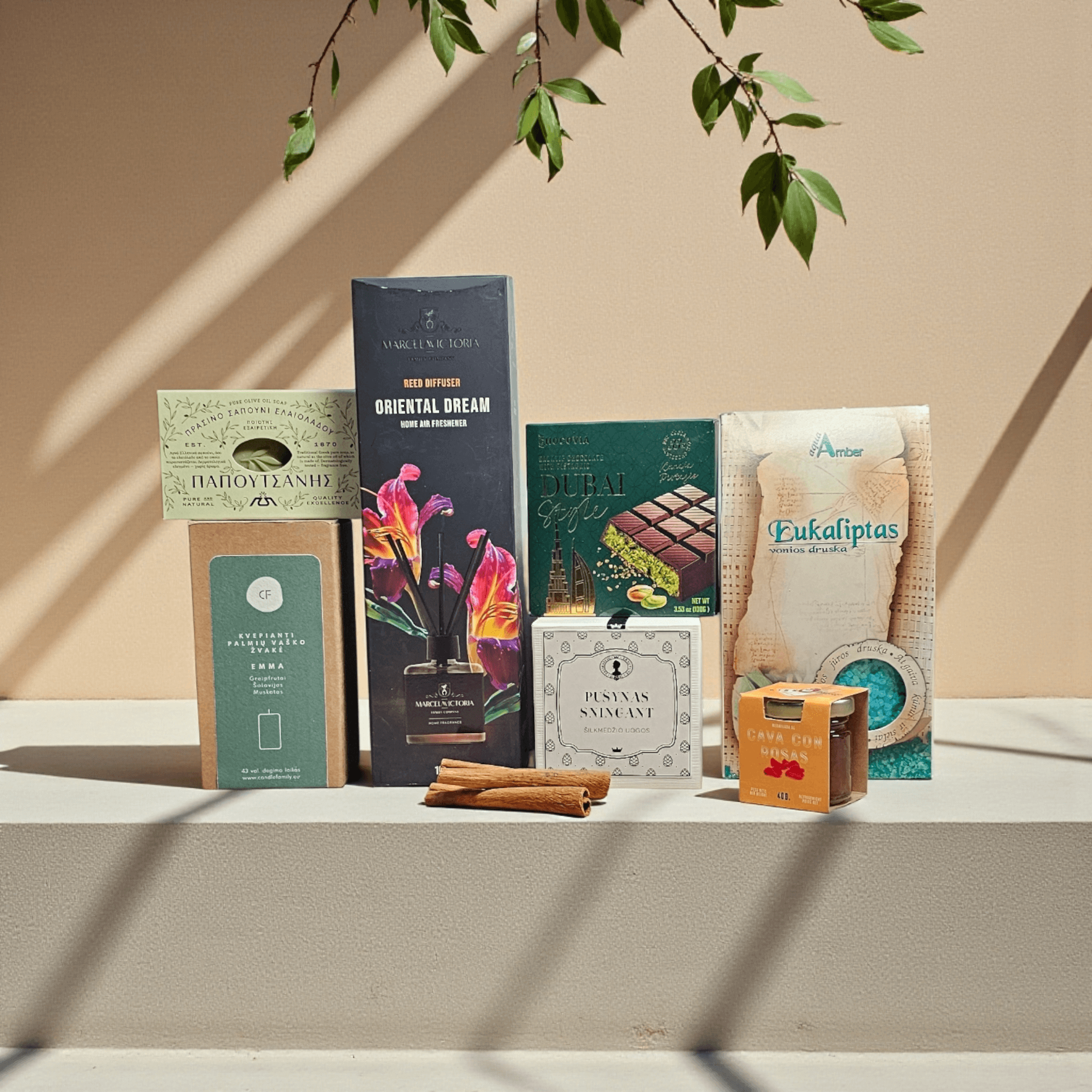 DORI gurmaniškas rinkinys su kvapų difuzoriumi, dubajaus šokoladu, vonios druska ir muilu, fotografuotas šiltame minimalistiniame fone su kabančiais žalumynais.