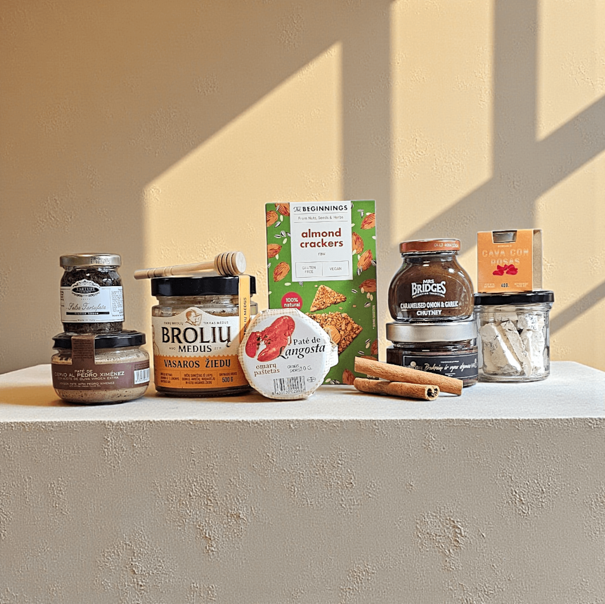 DORI gurmaniškas rinkinys su medumi, padažais, paštetais ir riešutų pagardais, fotografuotas šiltame fone su faktūrine platforma.