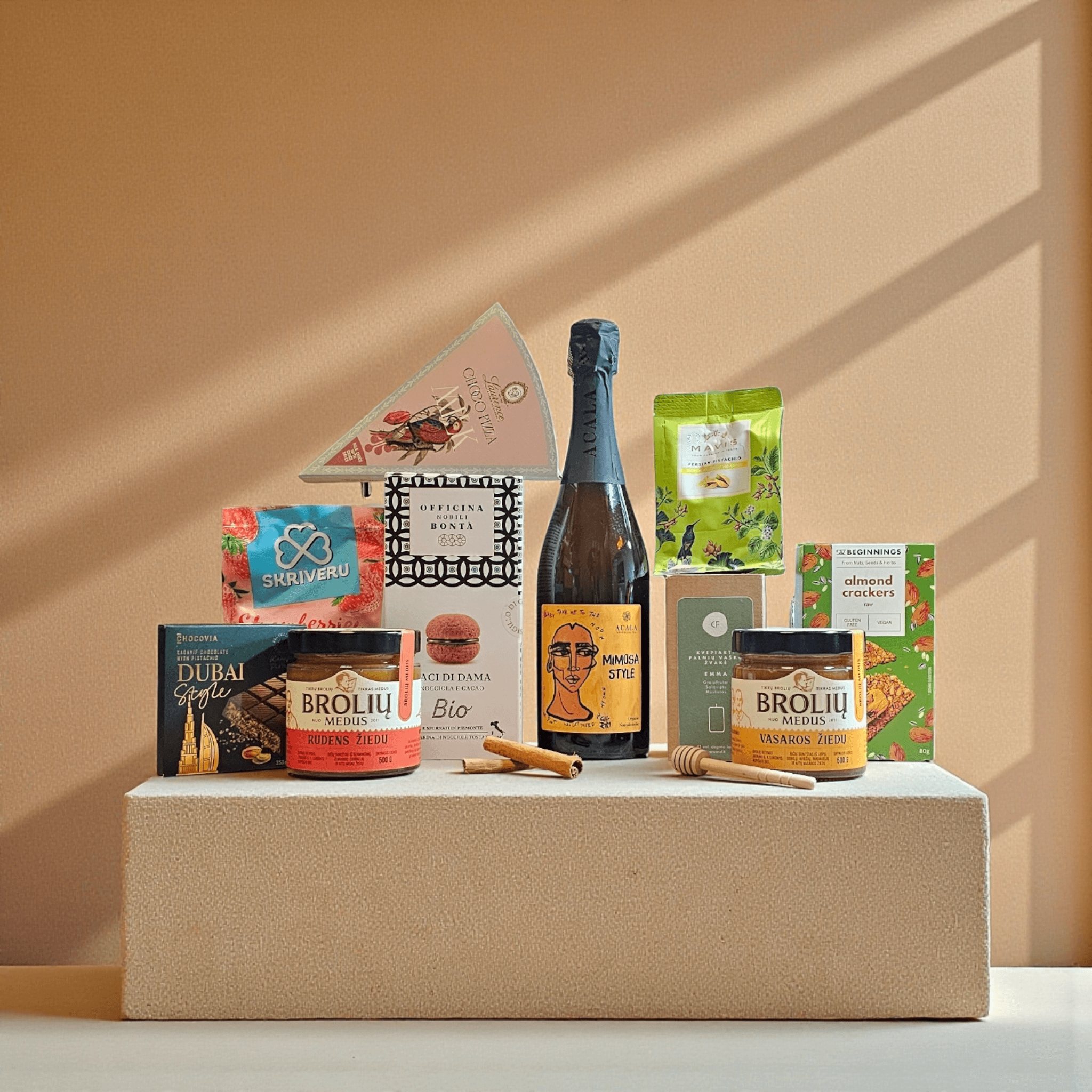 DORI gurmaniškas rinkinys su putojančiu vynu, šokoladu, skanėstais, arbata ir medumi, estetiškai sudėliotas ant faktūrinės platformos šiltame minimalistiniame fone.