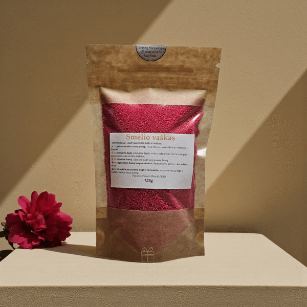 Raudonas smėlio vaškas 125 g kraft maišelyje su skaidriu langu, pastatytas ant minimalistinės šviesios pakylos, šiltame fone su švelniais šešėliais ir raudona gėle kompozicijoje.