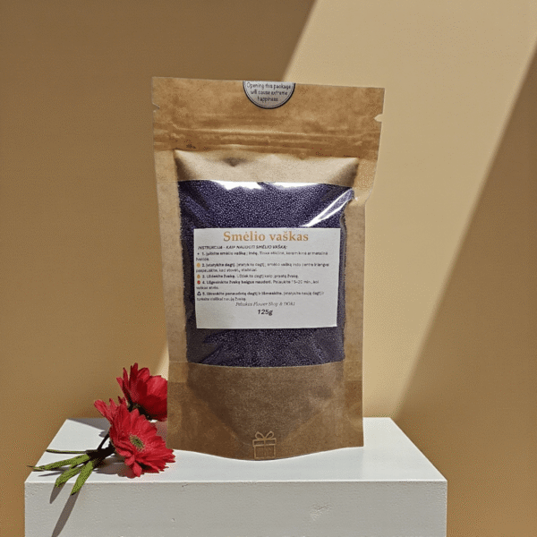 Purpurinis smėlio vaškas 125 g kraft maišelyje su skaidriu langu, pastatytas ant baltos minimalistinės pakylos, šiltame fone su krintančiais šešėliais ir raudonomis gėlėmis kompozicijoje.