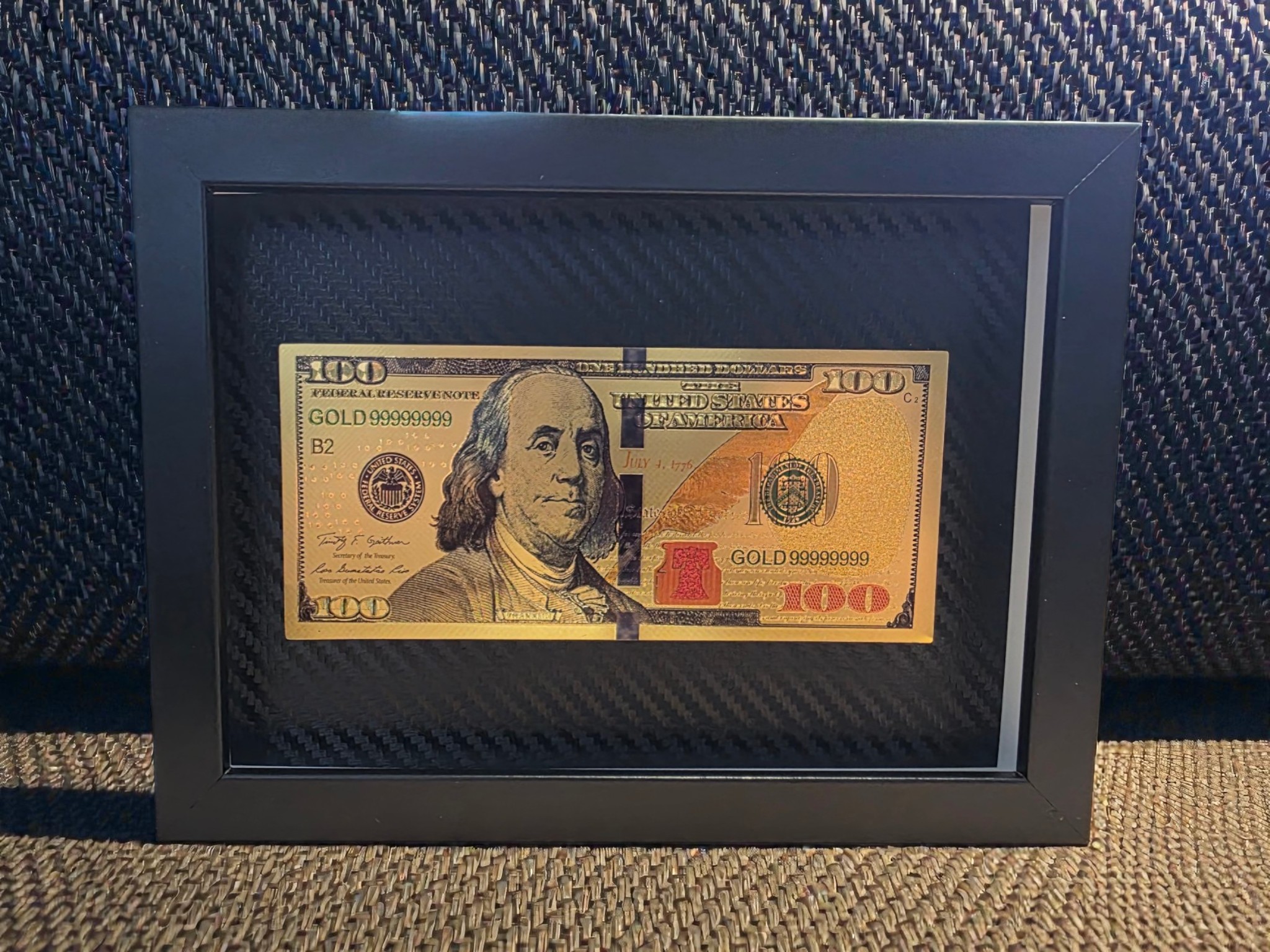 Auksinis 100 dolerių banknoto paveikslas juodame rėme su karbono tekstūros fonu