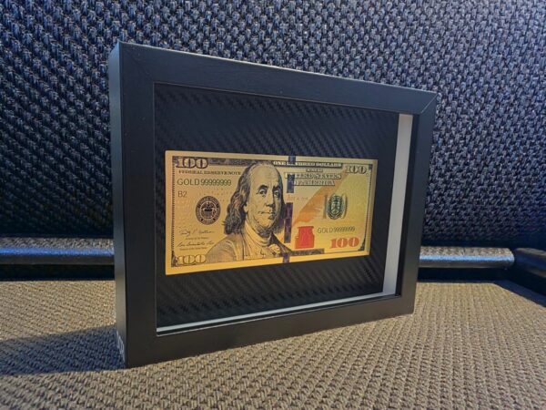 Auksinis 100 dolerių banknoto paveikslas juodame rėme su karbono tekstūros fonu