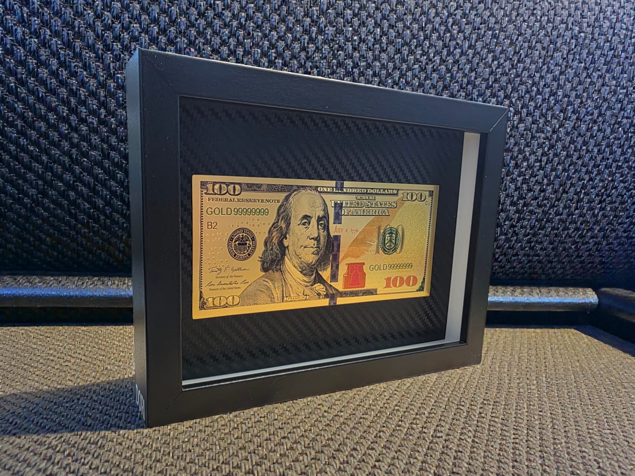 Auksinis 100 dolerių banknoto paveikslas juodame rėme su karbono tekstūros fonu